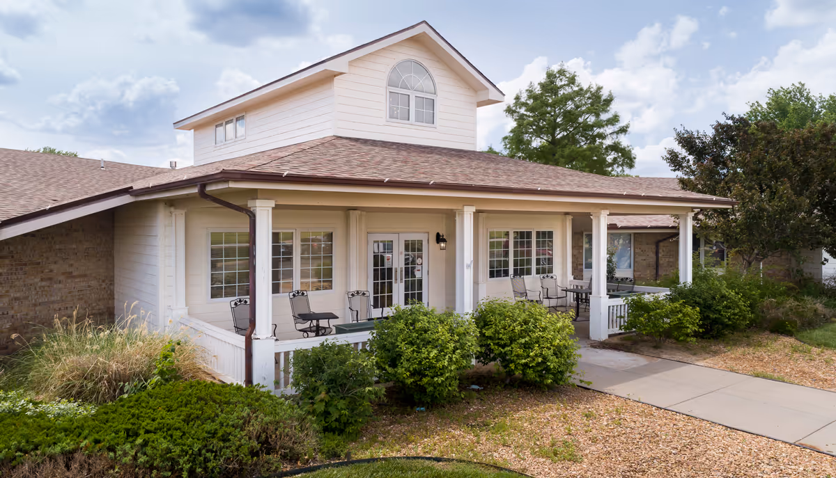Exterior view of a single-story building with a covered porch featuring several metal chairs and tables. The building has white siding, multiple windows, and a brown shingled roof. There are green bushes and landscaping in front of the porch, with a concrete walkway leading to the entrance. The sky is partly cloudy.