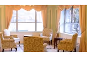 A bright living room with four patterned armchairs arranged around a small wooden table. Large windows with peach-colored curtains allow natural light to fill the space.