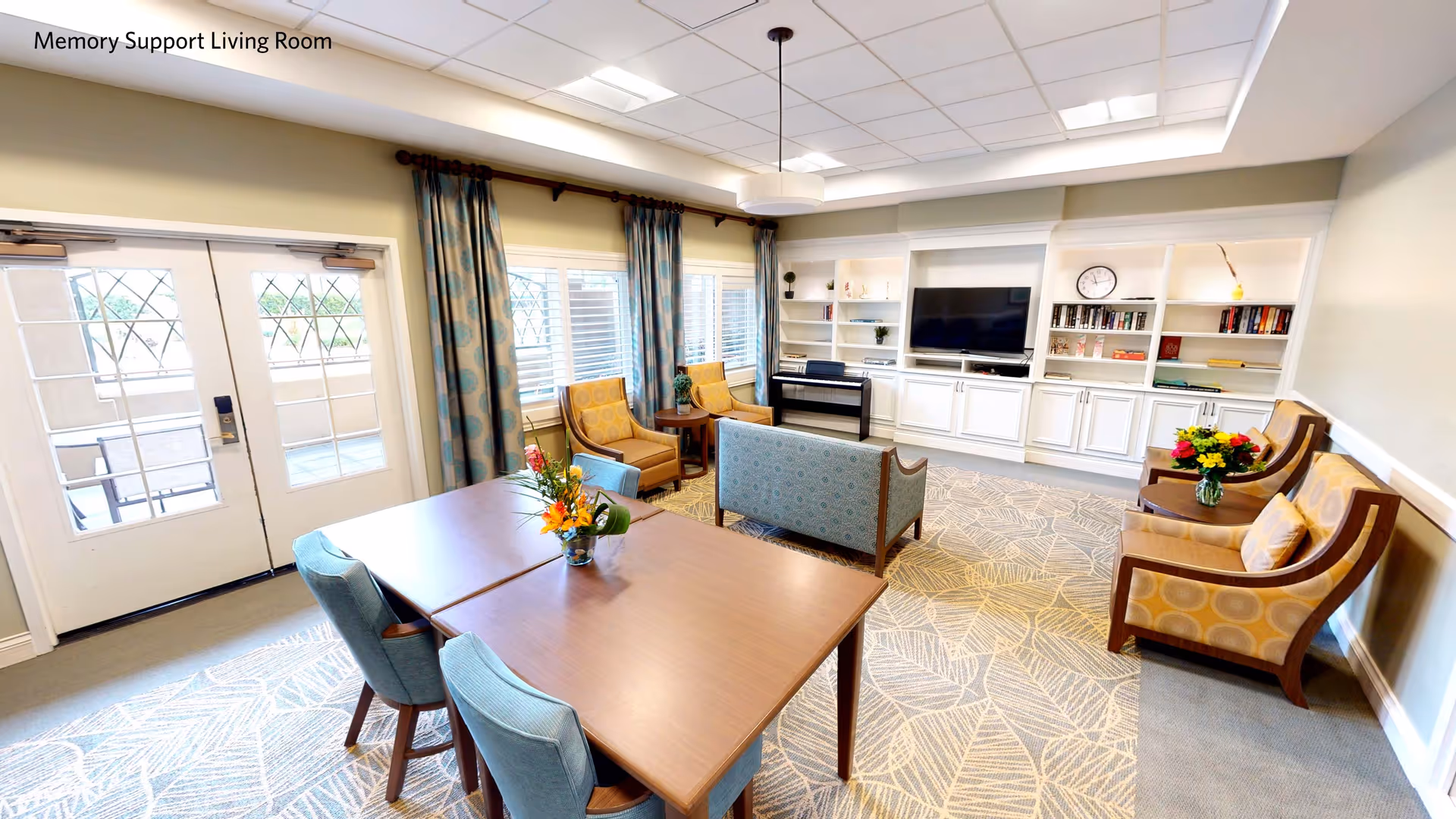 A bright and spacious memory support living room with a wooden table and four blue upholstered chairs in the foreground. The room features a patterned carpet, two yellow armchairs with a small round table between them, a blue patterned loveseat, and three additional yellow armchairs with a round table holding a vase of flowers. Built-in white shelves and cabinets line the back wall, with a flat-screen TV and a clock above. Large windows with blue patterned curtains and double glass doors let in natural light.
