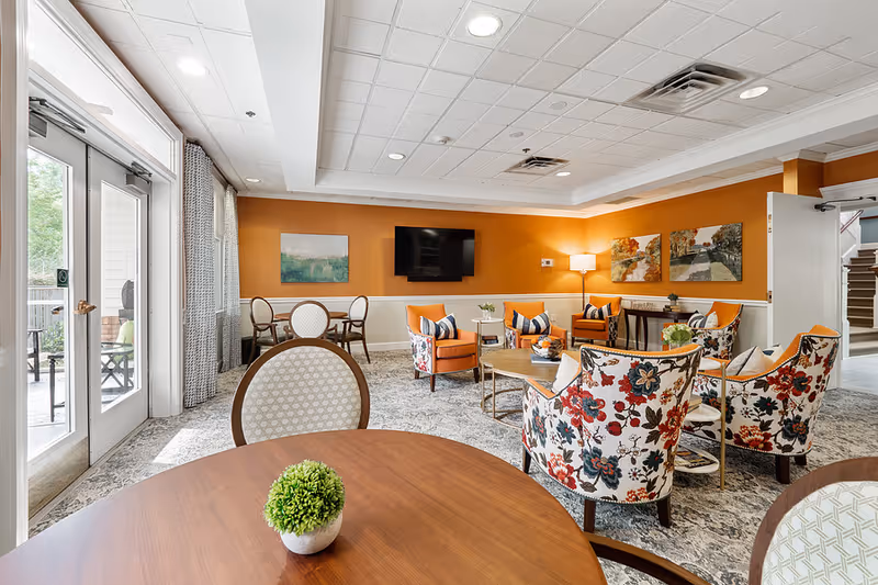 Bright communal living room with floral armchairs, orange accent wall, round tables, a wall-mounted TV and glass doors leading to a patio.