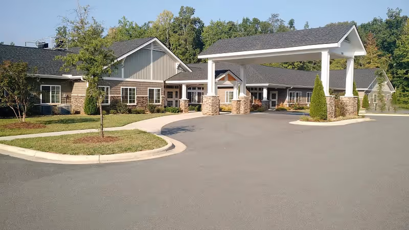 Exterior view of a single-story senior living facility building with a covered entrance supported by stone pillars, surrounded by a paved driveway and landscaped greenery with trees and shrubs.