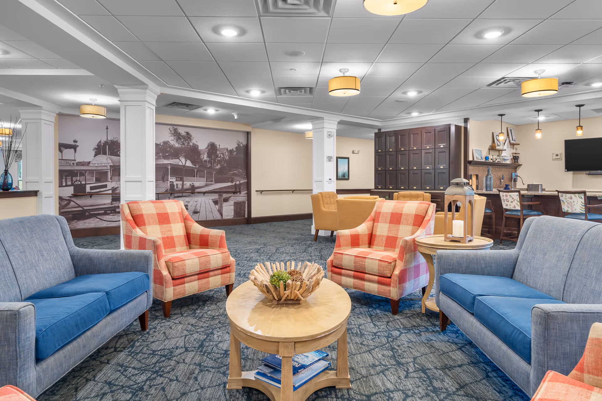 Comfortable common seating area with blue sofas, red plaid armchairs, tables, and a mural in a senior living facility.