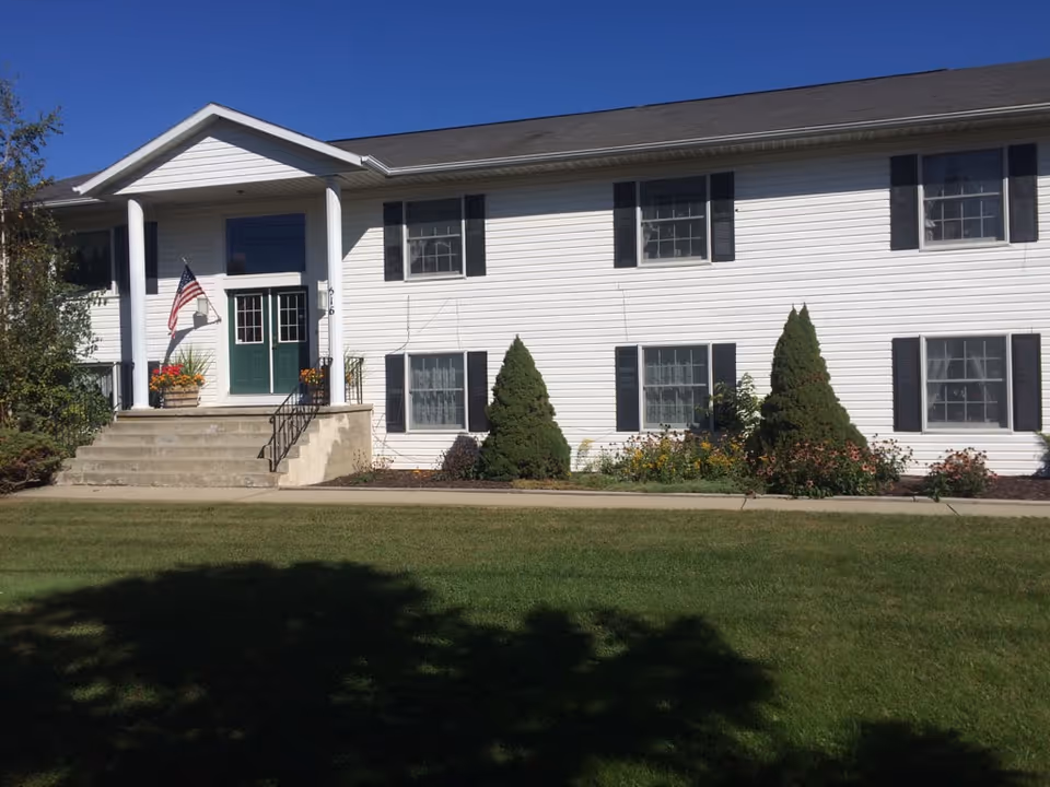 White two-story building with green double doors, front steps, an American flag, and a lawn.