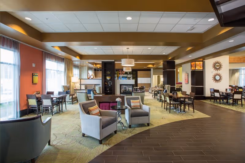A spacious and well-lit common area in a senior living facility featuring multiple seating arrangements including armchairs with decorative pillows, small round tables, and dining tables with chairs. The room has large windows with sheer curtains allowing natural light, warm-toned walls, and a mix of carpet and wood flooring. Decorative elements include wall art, a modern fireplace, and pendant lighting fixtures.