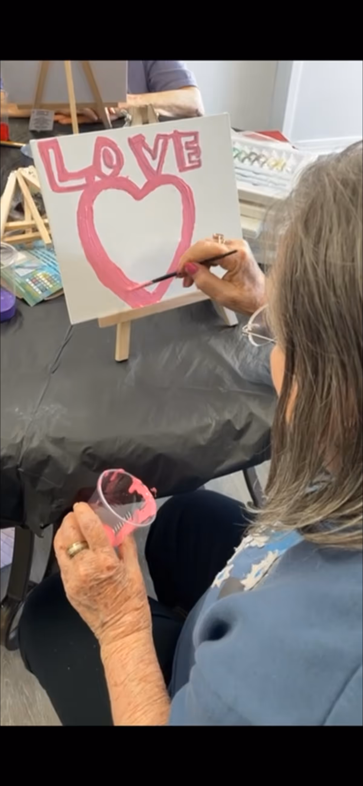 An elderly woman with gray hair is painting a canvas on an easel. She is painting a pink heart with the word 'LOVE' above it in pink letters. She holds a small cup of pink paint in one hand and a paintbrush in the other. The table is covered with a black plastic tablecloth and there are art supplies visible in the background.