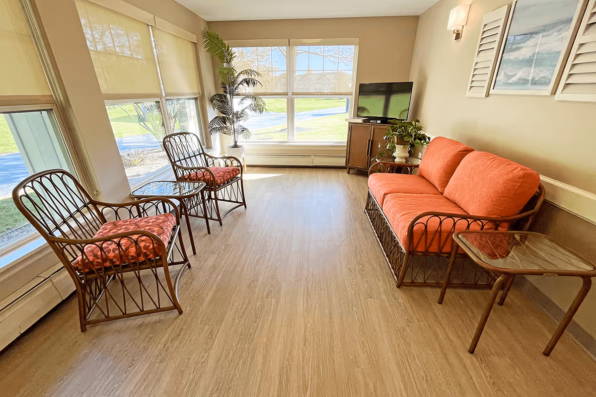 A bright and cozy sitting area with large windows letting in natural light. The room features a wicker sofa with orange cushions, two matching wicker chairs with patterned cushions, a glass-top side table, a small wooden cabinet with a flat-screen TV, a potted plant, and a framed painting on the beige wall.