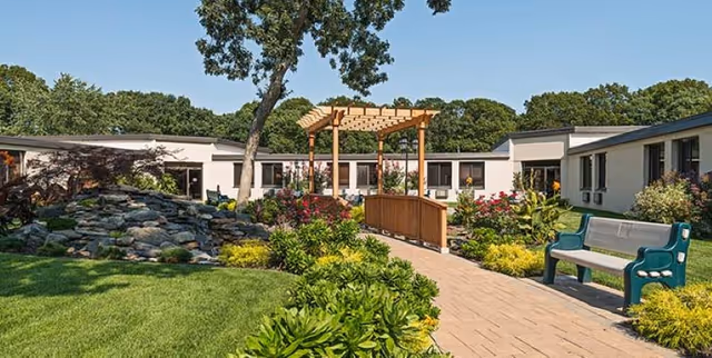 Outdoor garden area at The Arbors featuring a paved walkway, green benches, a wooden pergola, lush greenery, colorful flowers, and a rock water feature with trees and a single-story building in the background under a clear sky.