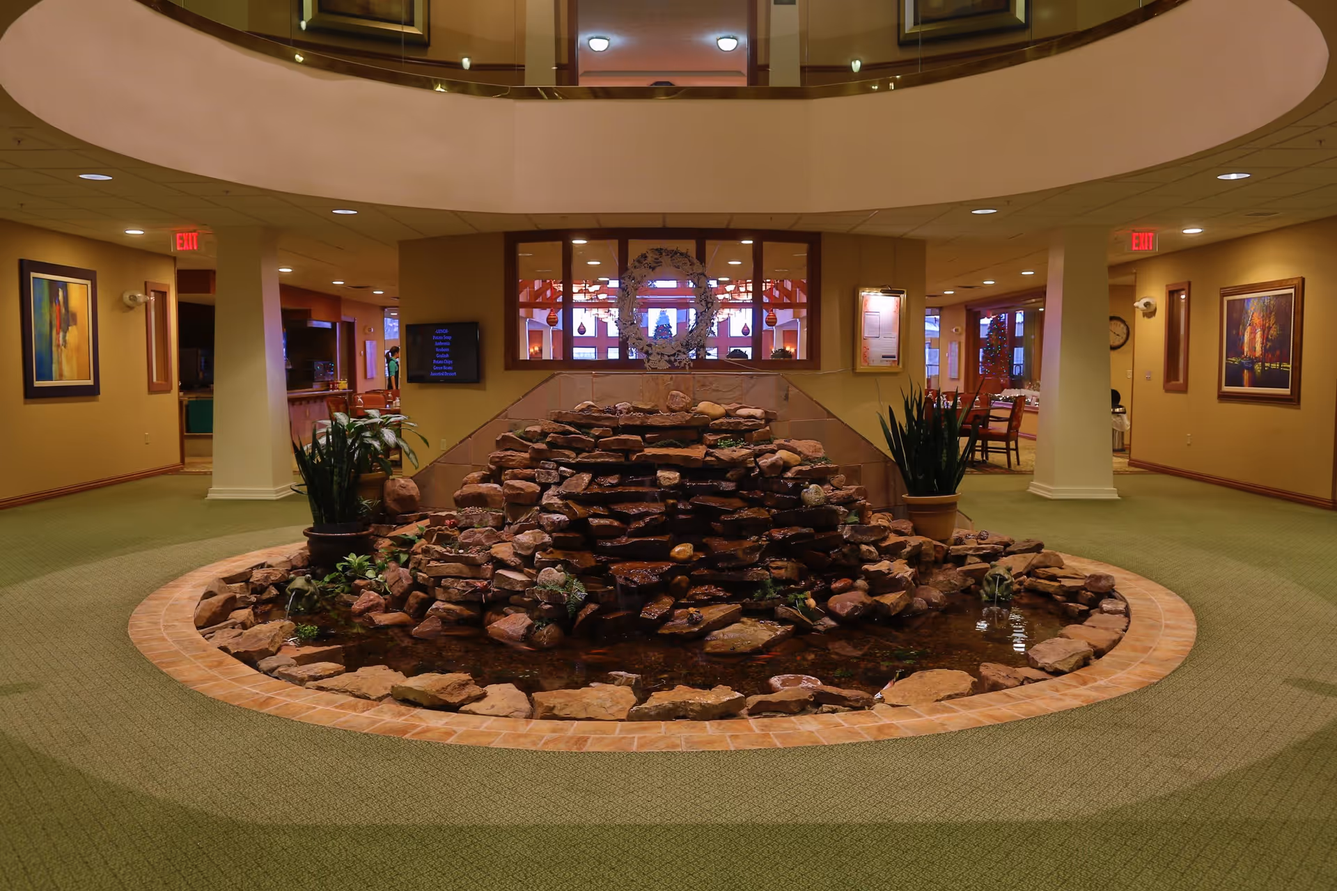 Indoor common area with a circular rock water feature in the center, surrounded by green carpet and beige walls. The space has columns, framed artwork on the walls, and visible seating areas in the background. The ceiling has recessed lighting and a circular opening above the water feature.