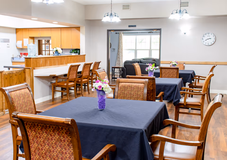 A dining area in a senior living facility with several tables covered in dark tablecloths, each adorned with a small vase of flowers. Wooden chairs with patterned upholstery surround the tables. In the background, there is a kitchen counter with bar stools and a living room area with sofas near a large window. The room has wooden flooring and is well-lit with ceiling lights and wall sconces.
