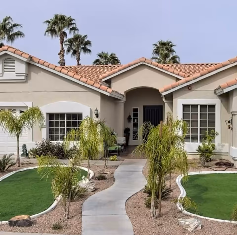 Front exterior view of a single-story building with a tiled roof, beige walls, and a curved concrete walkway leading to a dark front door. The landscape includes small palm trees, green grass patches, and desert-style gravel. Tall palm trees are visible in the background under a clear sky.