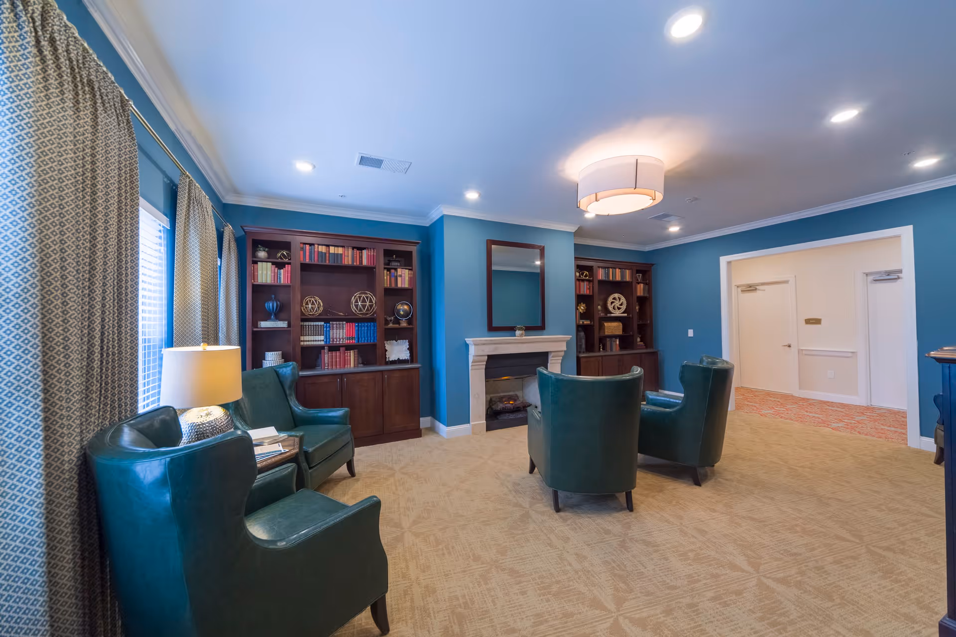 A cozy living room area with teal walls and beige carpet. The room features four dark green leather armchairs arranged around a white fireplace with a mirror above it. Two wooden bookshelves filled with books and decorative items flank the fireplace. Large windows with patterned curtains allow natural light into the room, and a modern ceiling light fixture illuminates the space.