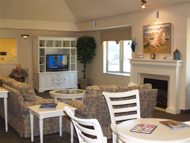 A cozy living room in an assisted living center featuring patterned sofas arranged around a glass coffee table, a white entertainment center with a TV, a potted plant, a fireplace with a painting above it, and a round table with chairs in the foreground. A person is seated on one of the sofas in the background.