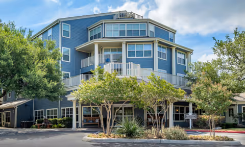 Exterior view of a multi-story blue retirement community building with white trim, surrounded by trees and landscaping under a partly cloudy sky.