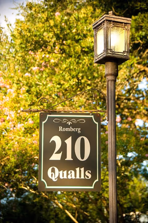 A decorative street lamp post with a hanging sign that reads 'Romberg 210 Qualls' surrounded by green trees in the background.