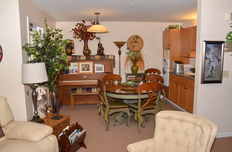Interior view of a senior living facility room featuring a round dining table with four wooden chairs, a wooden piano with framed photos and decorative items on top, a kitchen area with wooden cabinets and appliances, a beige armchair, a floor lamp, and various plants and wall decorations.