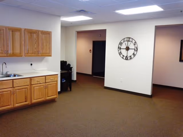Interior view of a room with wooden cabinets and a sink on the left side, beige walls, brown carpet, a large wall clock, and two open doorways leading to other rooms.