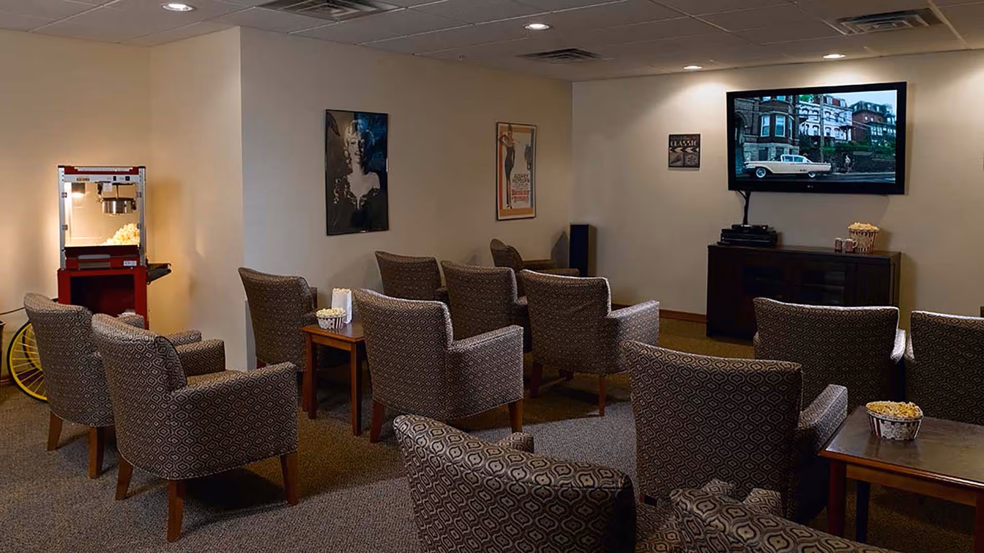 A cozy movie room with several patterned armchairs arranged in rows facing a wall-mounted flat-screen TV. There are small tables with popcorn buckets and a popcorn machine in the corner. The walls have framed artwork and movie posters.