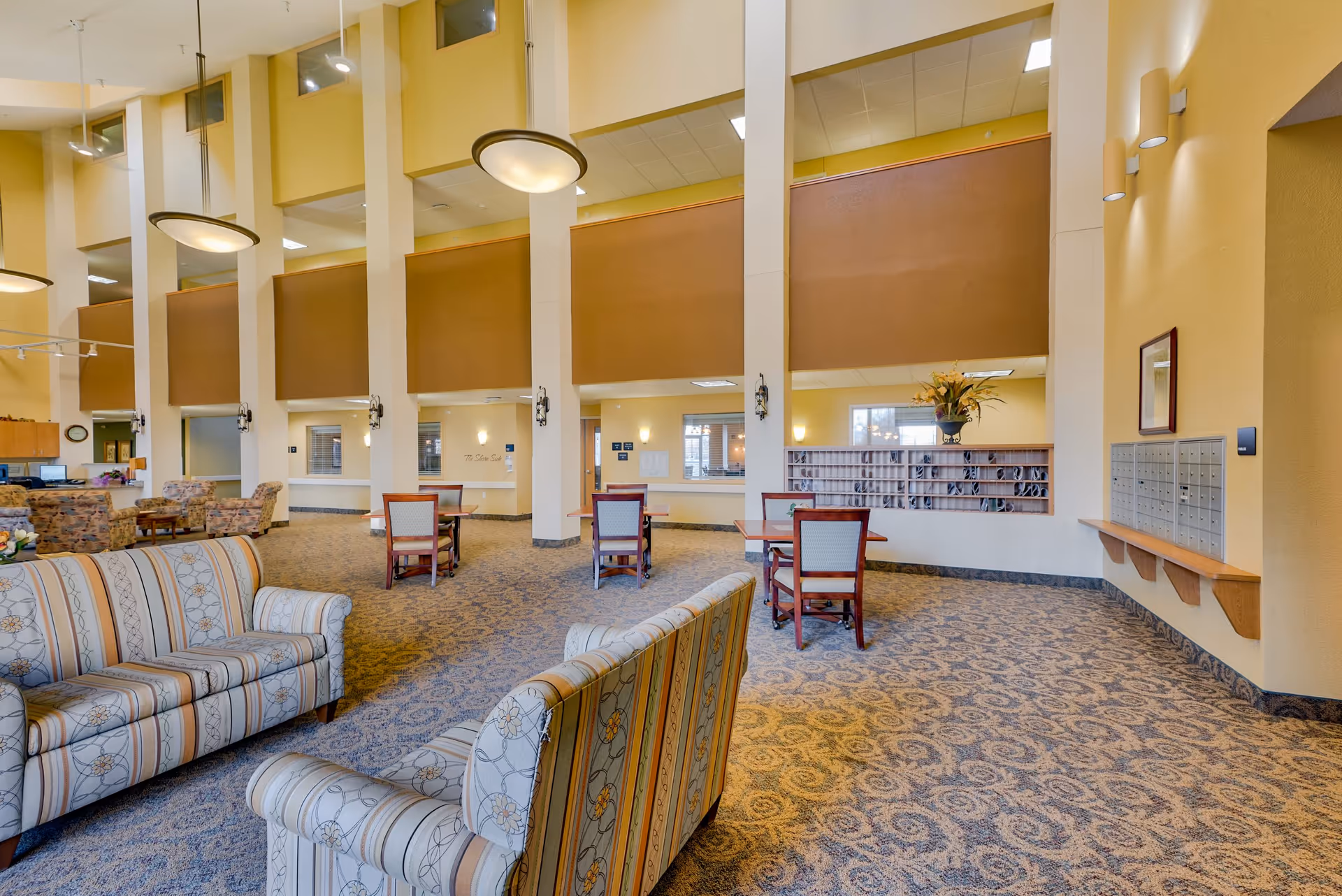 A spacious senior living facility common area with high ceilings and large hanging light fixtures. The room features patterned carpet, several upholstered sofas with striped and floral designs, wooden tables with chairs, and a wall with mailboxes. The walls are painted in warm yellow tones, and there are decorative elements such as a flower arrangement on a shelf and framed artwork.