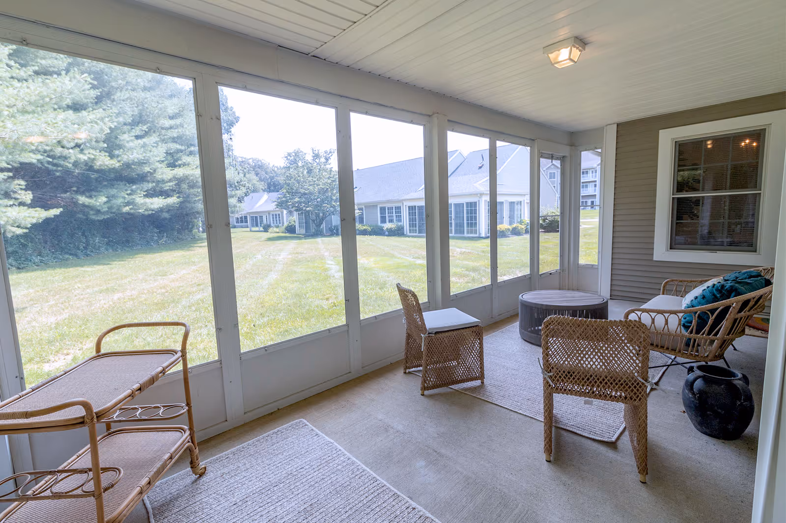 A screened-in porch area with wicker furniture including two chairs, a loveseat with cushions, a round coffee table, and a small serving cart. The porch overlooks a grassy lawn with trees and residential buildings in the background.