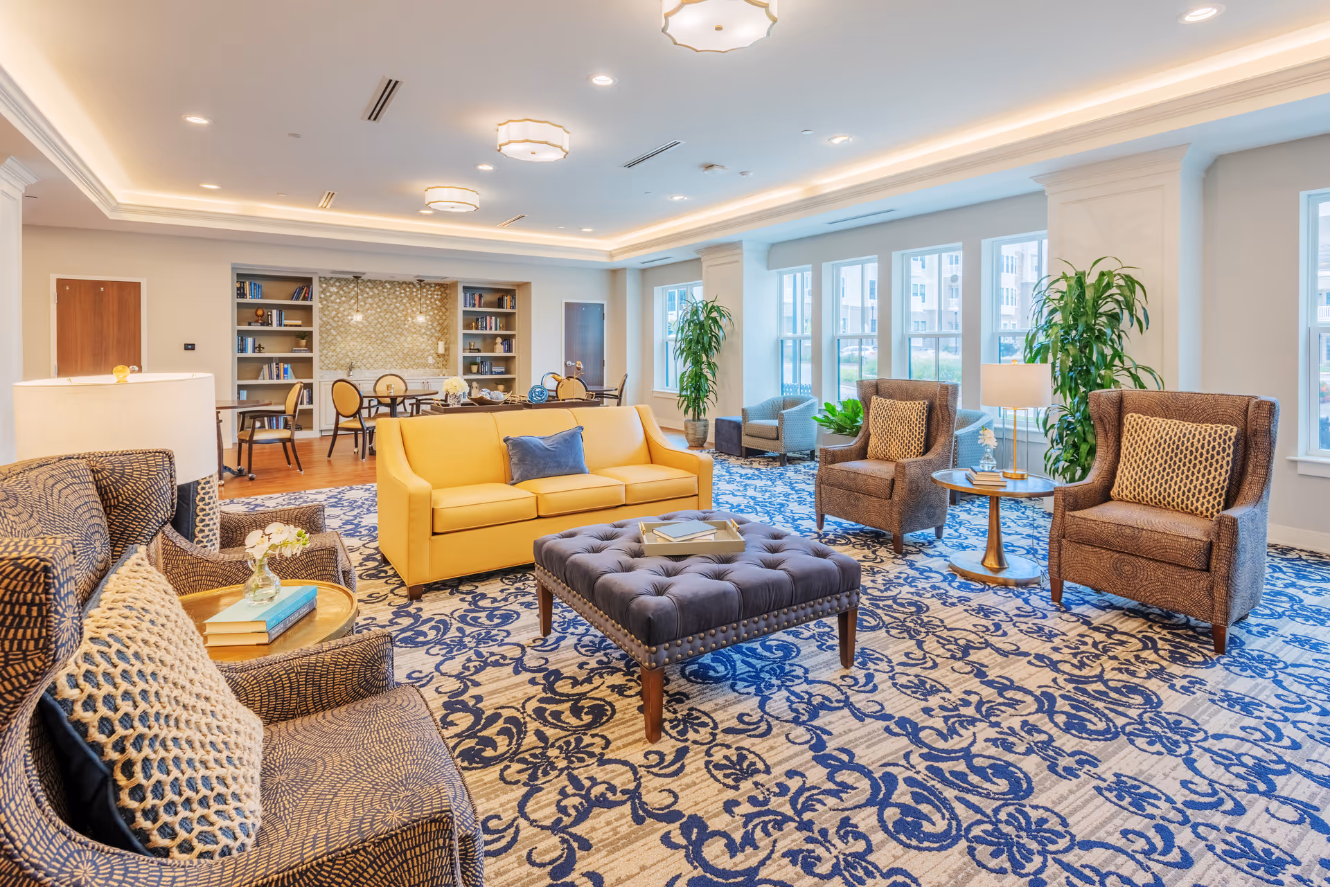A spacious and well-lit living room area with large windows allowing natural light to fill the space. The room features a yellow sofa, several patterned armchairs with cushions, a tufted ottoman coffee table, side tables with lamps, and decorative plants. In the background, there are bookshelves and a dining table with chairs. The room has a blue and beige patterned carpet and recessed ceiling lighting.