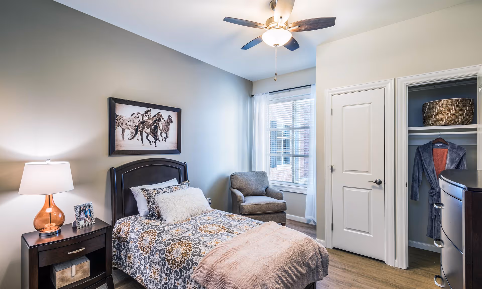 Furnished bedroom with a single bed, nightstand and lamp, armchair by a window, ceiling fan, and an open closet.