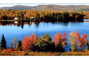 A scenic view of a calm lake surrounded by trees with autumn foliage in vibrant red, orange, and yellow colors, with hills in the background under a clear sky.