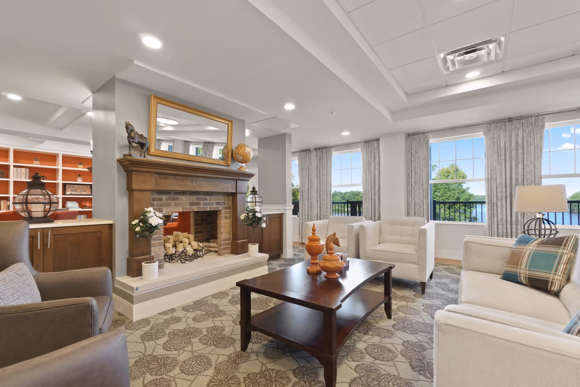 A bright and spacious senior living facility common area featuring a cozy brick fireplace with candles and decorative items on the mantel, including a globe and horse figurine. The room has large windows with gray curtains offering a view of greenery and water outside. Comfortable seating includes white armchairs and a sofa with plaid pillows, arranged around a dark wooden coffee table with decorative orange vases. The carpet has a floral pattern, and the walls are painted light gray with recessed lighting in the ceiling.