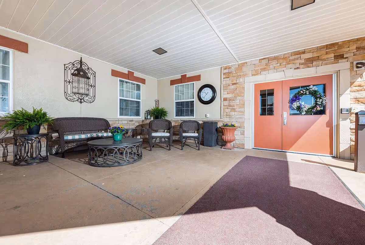 Covered outdoor seating area with wicker chairs, a wicker loveseat, a round coffee table, potted plants, a wall clock, and double doors decorated with a floral wreath.
