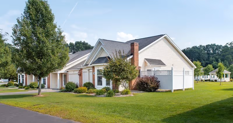 Single-story beige residential building with a white fence, manicured lawn and trees under a blue sky.