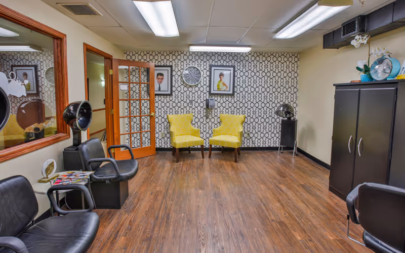 Interior of a hair salon or beauty parlor with wooden flooring, two yellow patterned chairs against a patterned wall with a clock and framed photos, black salon chairs, hair dryers, a black cabinet with decorative items, and a glass-paneled wooden door.