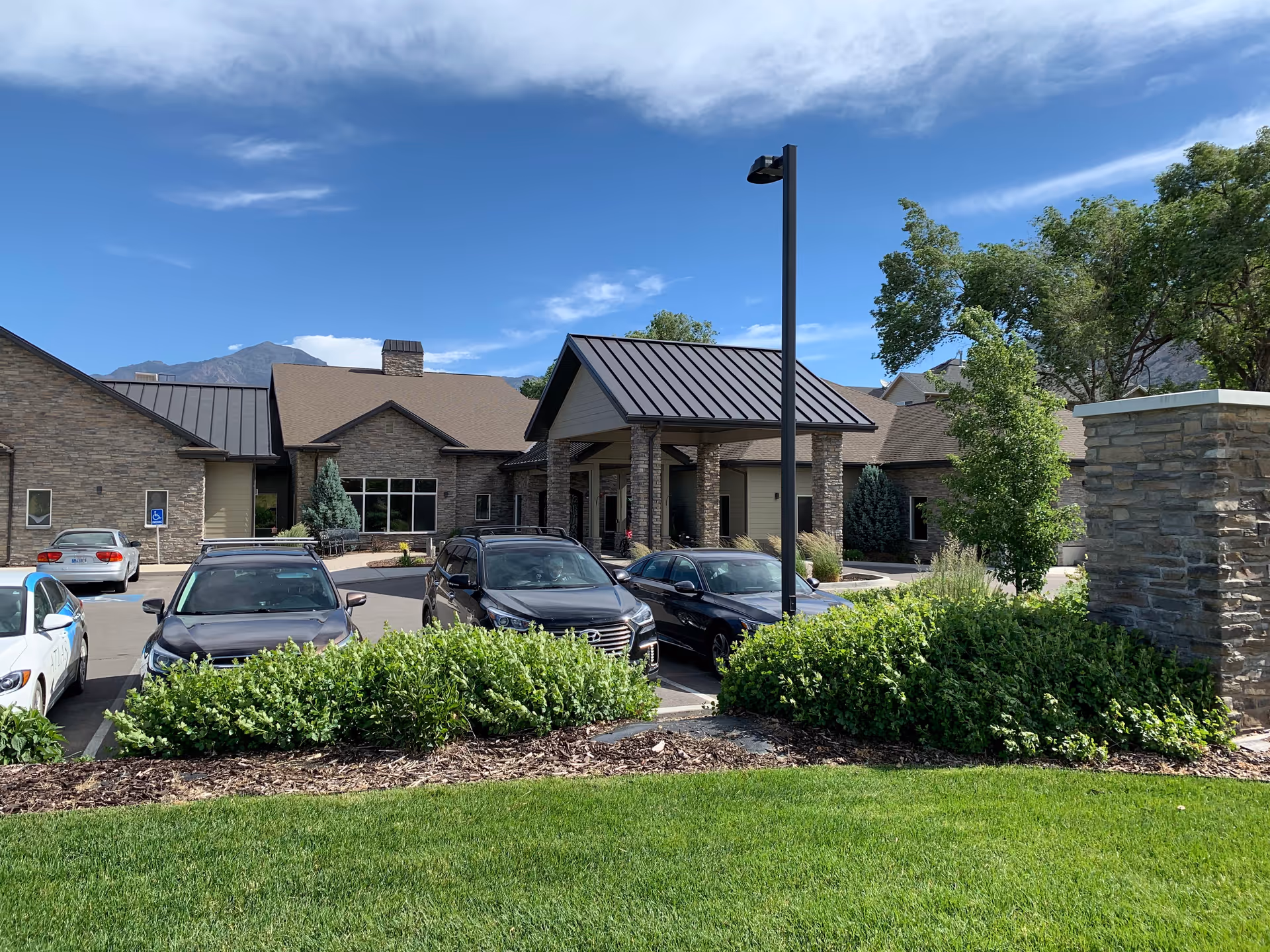 Exterior view of a single-story assisted living facility building with stone and beige siding, a covered entrance supported by stone pillars, several parked cars in front, green bushes, and a well-maintained lawn under a blue sky with some clouds.