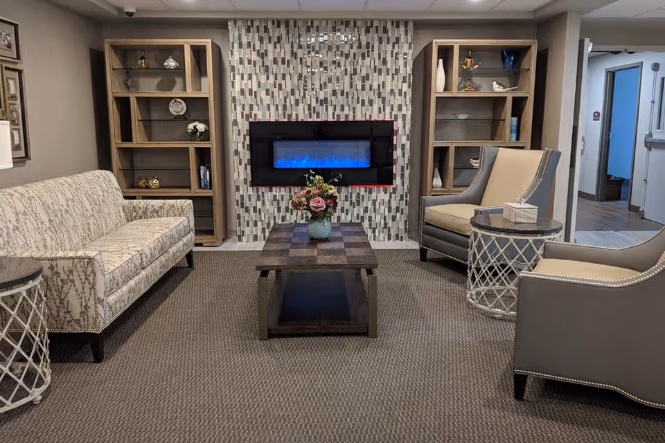 A cozy living room area with a patterned sofa on the left, two beige and gray armchairs on the right, and a wooden coffee table with a flower vase in the center. Behind the coffee table is a modern electric fireplace mounted on a tiled wall, flanked by two wooden shelving units with decorative items. The room has carpeted flooring and a doorway leading to another area.