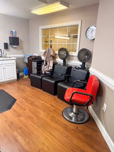 Salon area in an assisted living facility with three black hair-dryer chairs, a red styling chair, a sink, and cabinets.