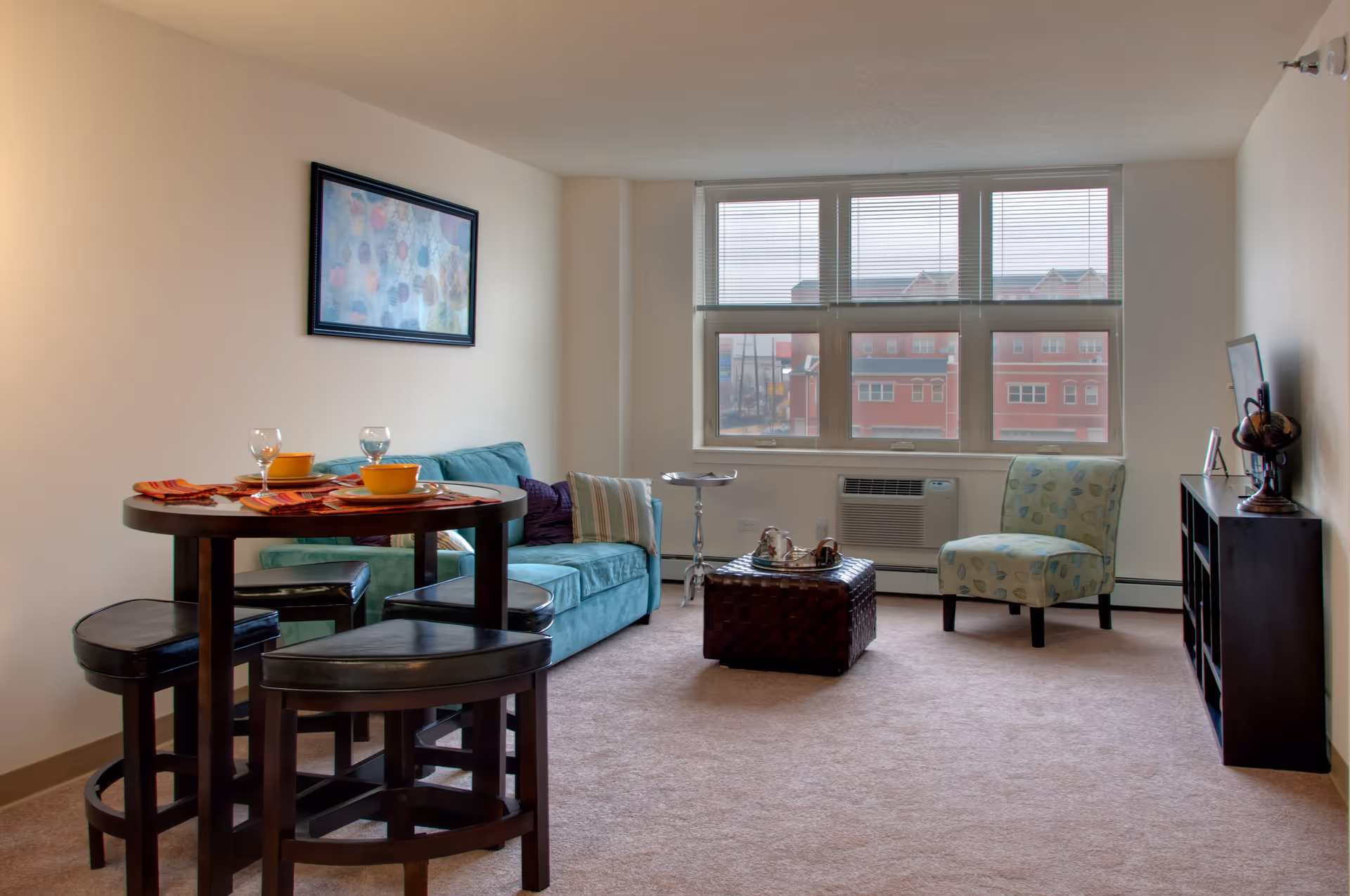 A bright living room with a large window showing a view of red buildings outside. The room features a teal sofa with pillows, a patterned armchair, a dark wooden coffee table with a decorative tray, a small round dining table with four black cushioned stools, and a black shelving unit with a globe and framed photo. A colorful abstract painting hangs on the wall above the sofa.