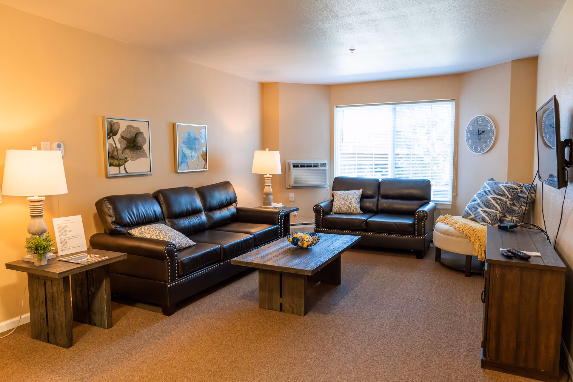 A cozy living room with two dark brown leather sofas, a wooden coffee table, and a side table with a lamp and small plant. There are two framed floral artworks on the wall, a window with blinds, an air conditioning unit, a wall clock, and a TV mounted on the wall opposite the sofas. A cushioned chair with pillows and a yellow throw blanket is also visible.