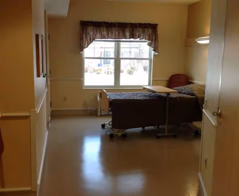 A hospital-style adjustable bed with a tray table in a beige, windowed patient room.