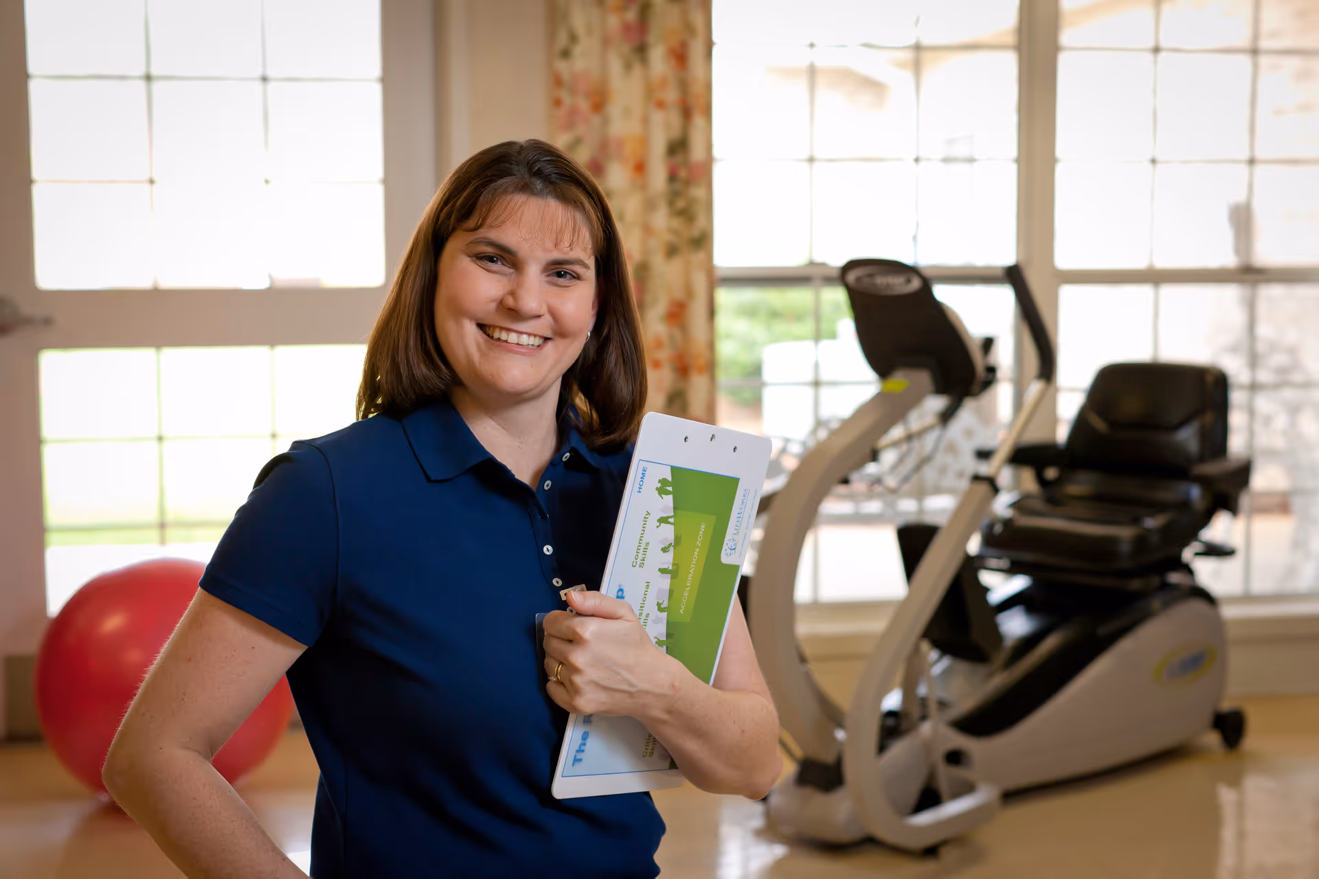 A smiling woman in a navy blue polo shirt holding a clipboard stands in a well-lit exercise room with large windows. Behind her, there is exercise equipment including a stationary recumbent bike and a large red exercise ball.