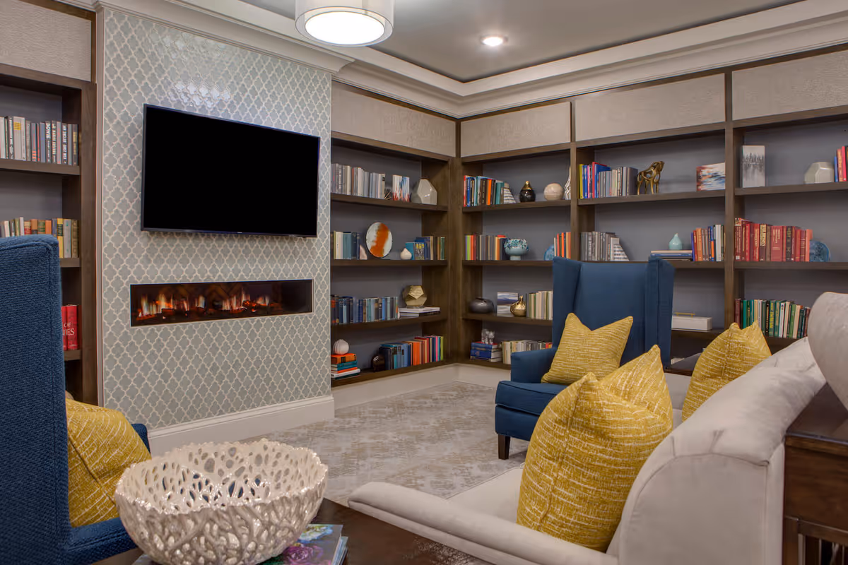 Cozy living room area with built-in bookshelves filled with books and decorative items, a wall-mounted flat screen TV above a modern electric fireplace, blue armchairs with yellow pillows, and a beige sofa with yellow pillows, all arranged on a patterned rug under a ceiling light.