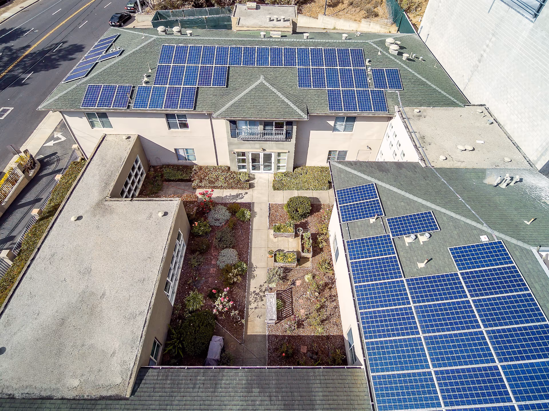Aerial view of Mission Villa Senior Living showing a courtyard garden surrounded by buildings with green roofs covered in solar panels. The courtyard has pathways, plants, and a bench. A street with parked cars is visible on the left side.
