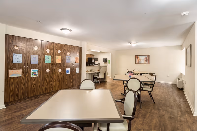 Communal activity/dining room with tables and chairs, a wall of wooden cabinets decorated with artwork, and a small service counter area.