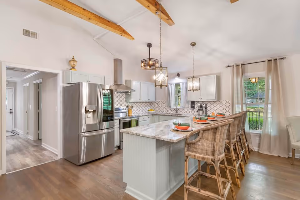 Bright and modern kitchen with a large marble island countertop surrounded by four wicker bar stools. The kitchen features stainless steel appliances including a refrigerator and stove, white cabinets, a patterned tile backsplash, and two pendant lights hanging from a vaulted ceiling with exposed wooden beams. There are windows with beige curtains letting in natural light and a view of greenery outside.