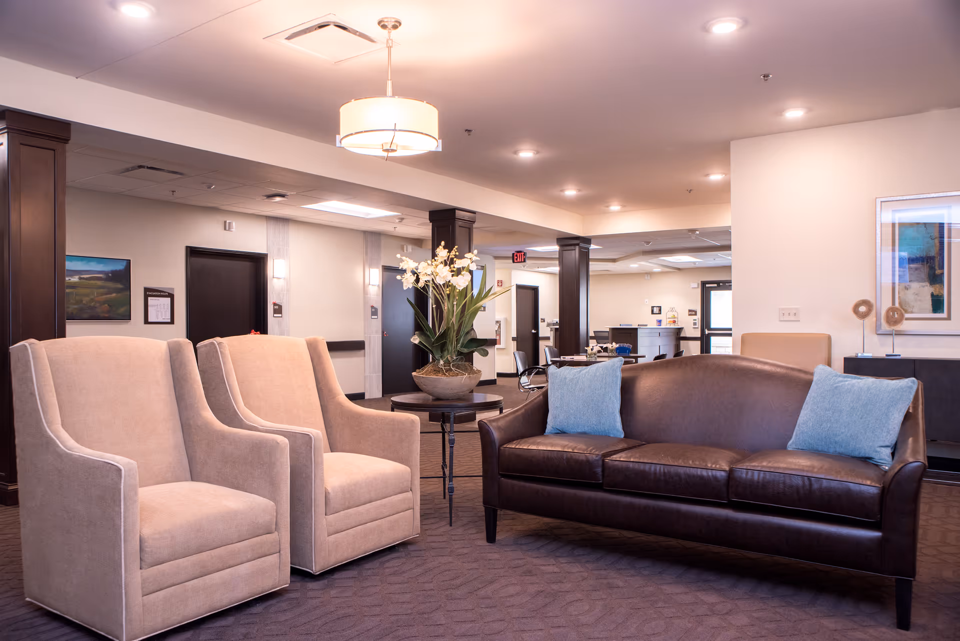 A senior living facility common area with two beige armchairs and a dark brown leather sofa with blue cushions arranged around a small round table holding a potted plant with white flowers. The room has soft lighting, dark wooden columns, and light-colored walls with framed artwork.