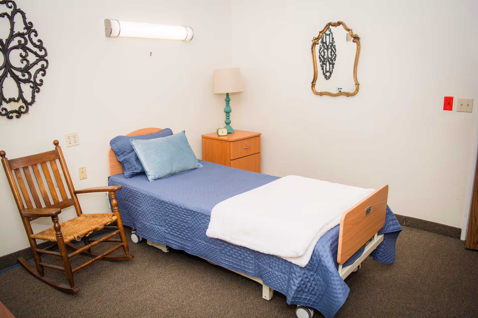 A small nursing facility bedroom with a single bed dressed in blue bedding, a wooden rocking chair, a nightstand with a lamp, and decorative wall mirrors.