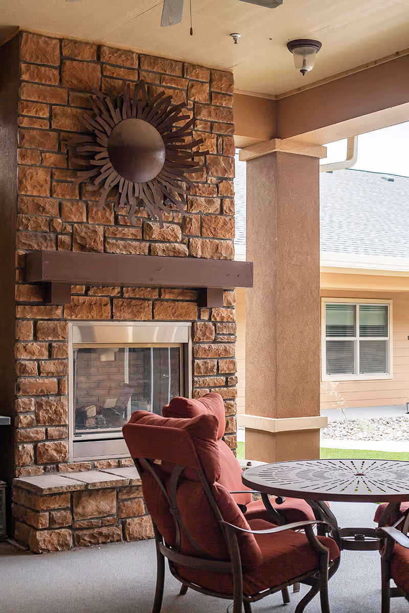 Covered outdoor seating area with a stone fireplace, metal sunburst wall art, and red cushioned chairs around a round table.