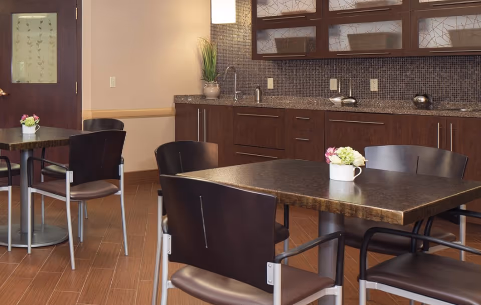 A small dining area with dark wood tables and chairs, each table decorated with a small white vase containing flowers. The background features a countertop with a sink, cabinets with glass doors, and a tiled backsplash. The floor has a wood-like tile pattern.