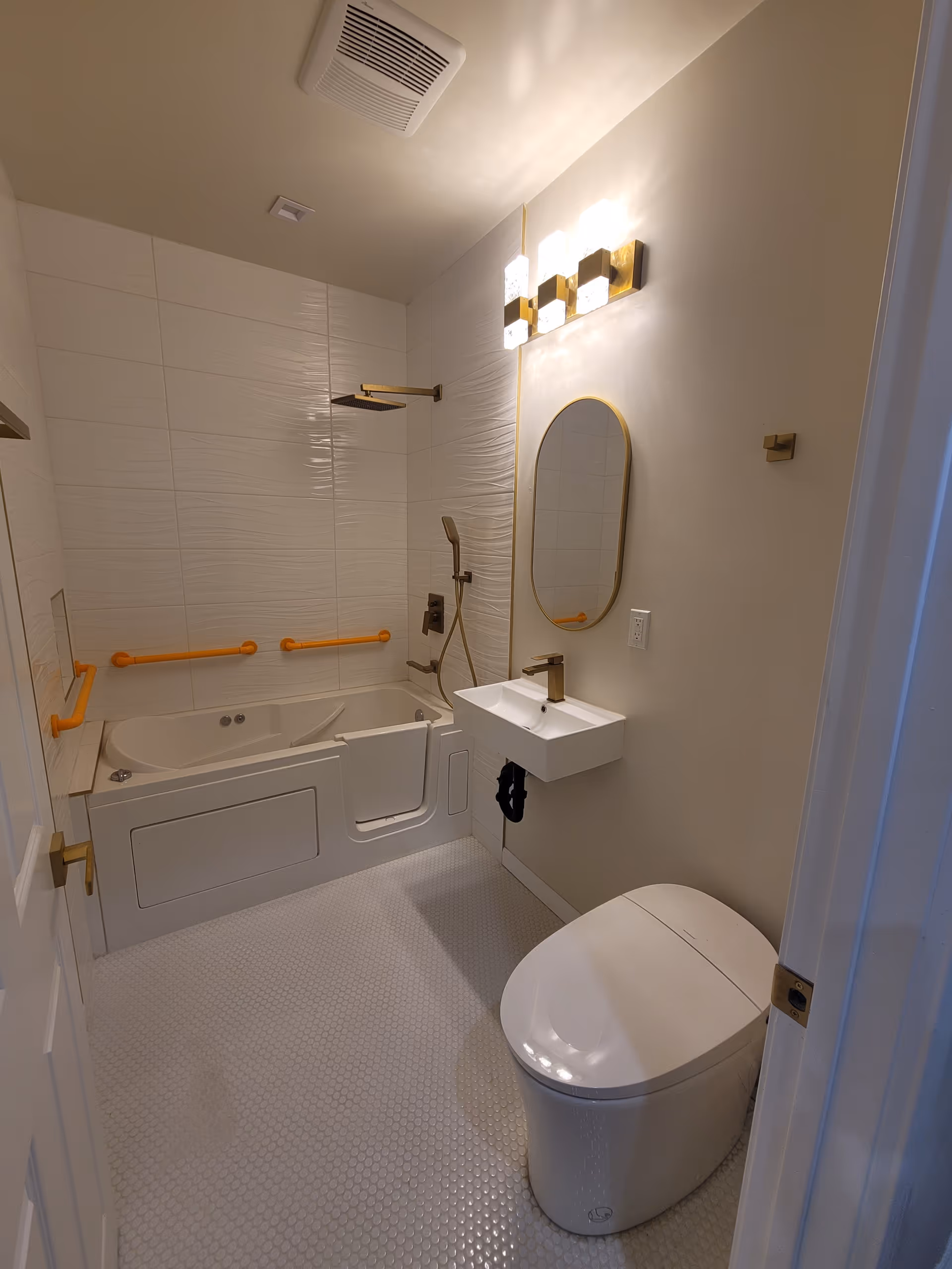 A modern bathroom featuring a walk-in bathtub with orange safety grab bars, a wall-mounted showerhead, a small white wall-mounted sink with a gold faucet, an oval mirror with a gold frame, a white toilet, and a light fixture with three bulbs above the mirror. The floor has small white hexagonal tiles and the walls are light-colored.