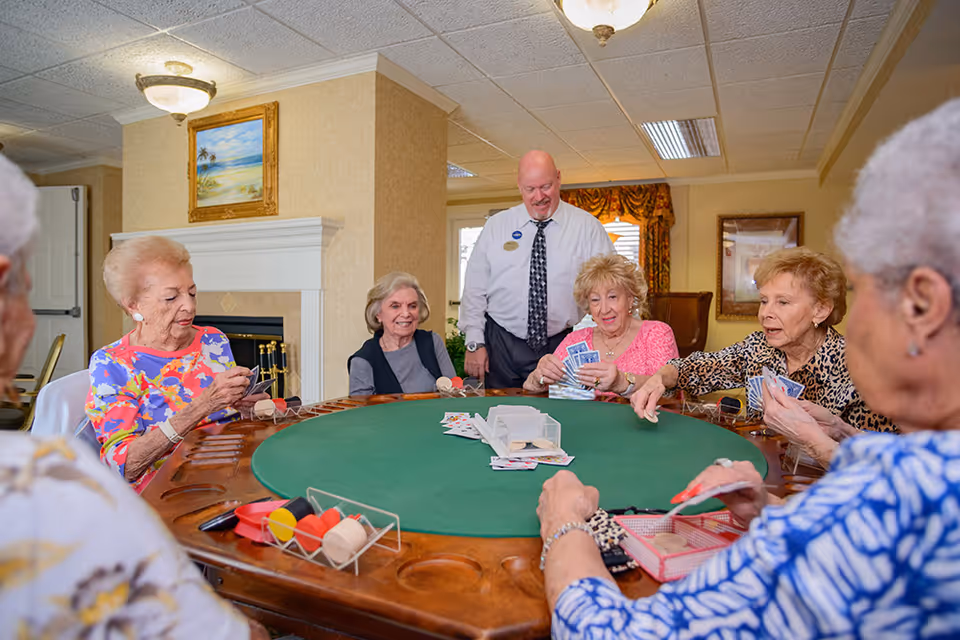 A group of elderly women sitting around a large round green felt card table playing cards, with a man standing behind them smiling. The room has beige walls, a fireplace with a painting above it, and a window with patterned curtains.