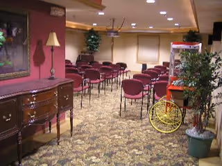 Interior room with rows of maroon chairs arranged facing a wall, a wooden sideboard with a lamp and framed picture on the left, a popcorn machine on the right, and potted plants placed around the room.