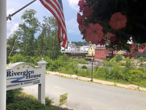 View of a street with a Riverglen House sign for independent and assisted living on the left side, an American flag above it, pink flowers hanging on the right, and a scenic background featuring trees, a river, and colorful buildings across the river under a partly cloudy sky.
