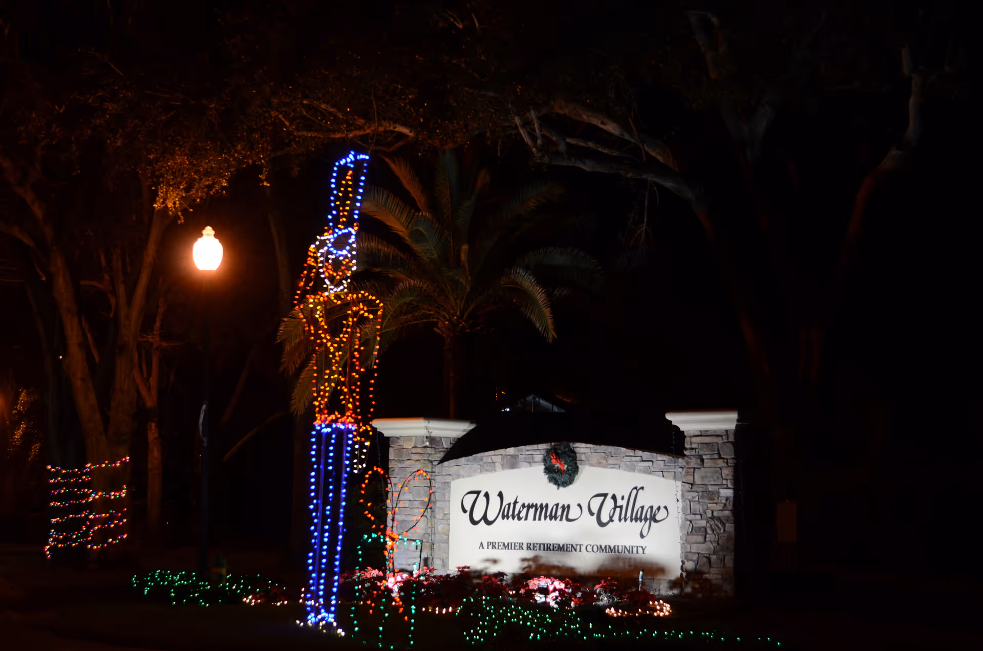 Nighttime view of the Waterman Village retirement community entrance sign decorated with holiday lights, including a large illuminated nutcracker figure and surrounding festive greenery and lights.